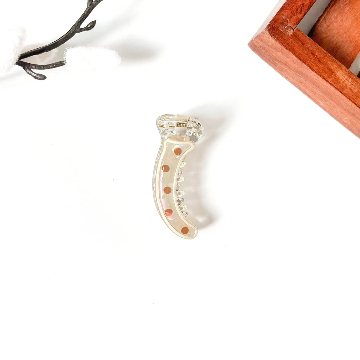 A curved clear resin hair mini banana clip with orange rhinestone accents, displayed on a clean white background with decorative props.