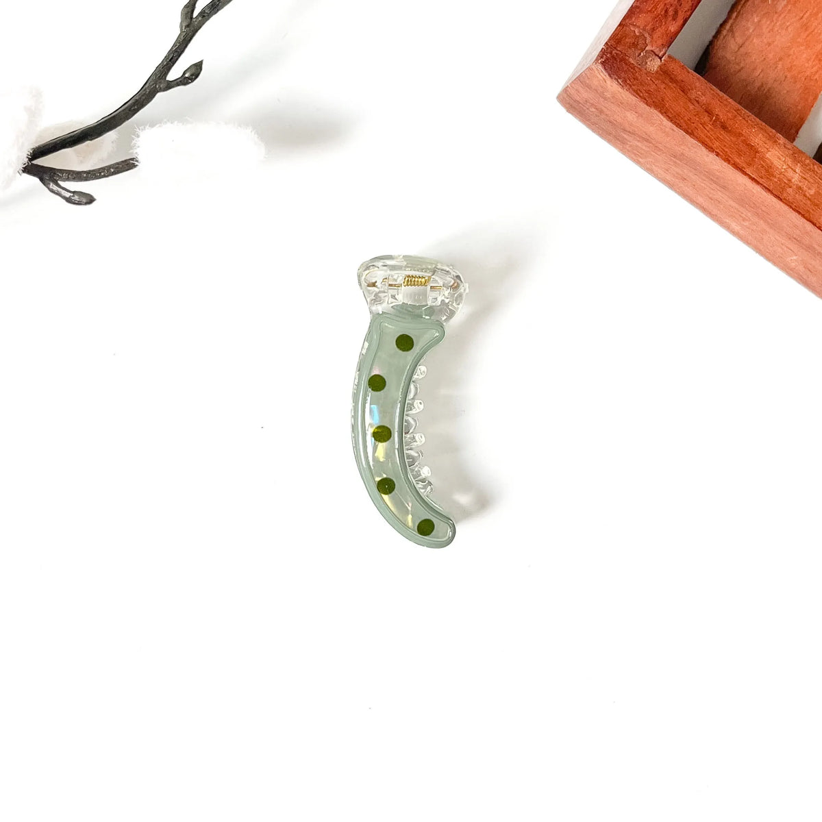 A small crescent-shaped clear resin or glass mini banana clip with green polka dots and a clear faceted top, photographed on a white background with partial decorative props at the corners.