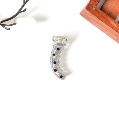 A curved clear acrylic hair mini banana clip with iridescent rhinestones and dark blue rhinestones, displayed on a clean white background with decorative props.