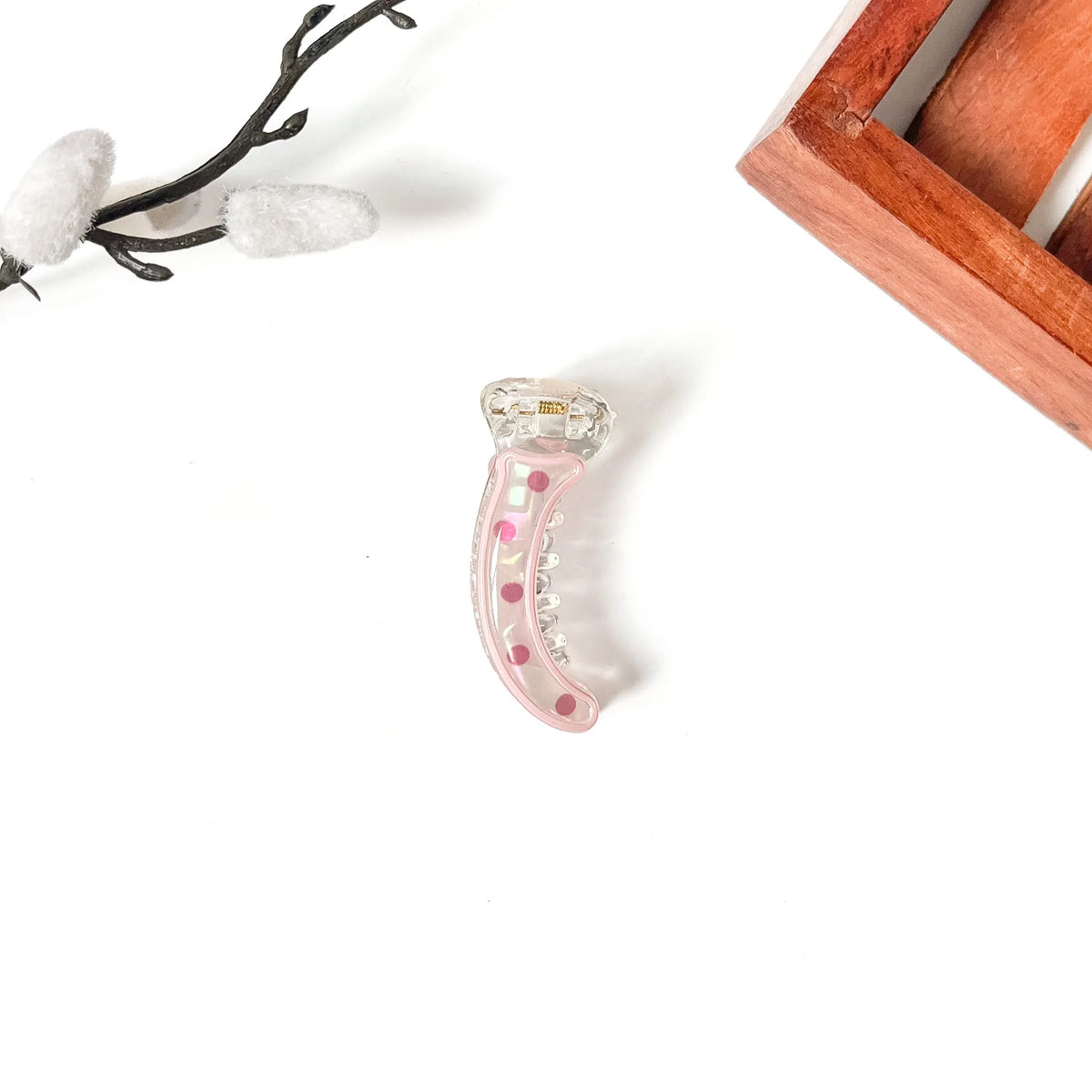 A curved claw-style mini banana clip made of transparent acrylic with pink polka dots and a small gold logo, displayed on a clean white background with decorative props.
