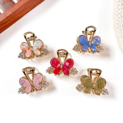 Five resin butterfly metal claw clips in coral, mauve pink, hot pink, sapphire blue, and khaki olive with gold-tone frames and crystal bead accents, arranged flat on cream background.