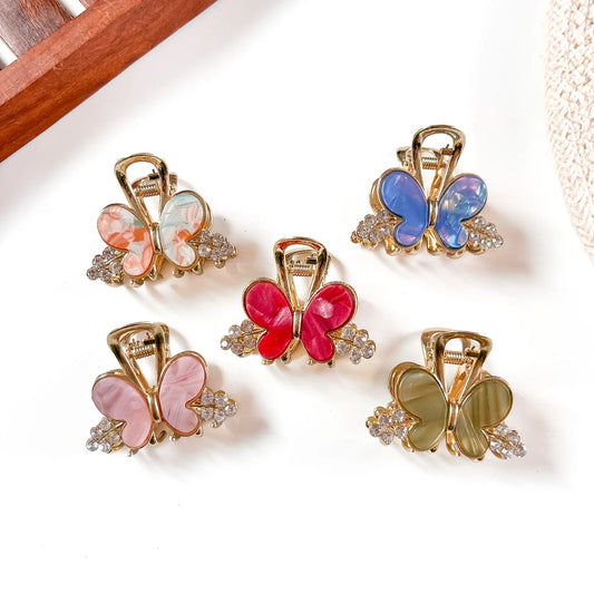Five resin butterfly metal claw clips in coral, mauve pink, hot pink, sapphire blue, and khaki olive with gold-tone frames and crystal bead accents, arranged flat on cream background.