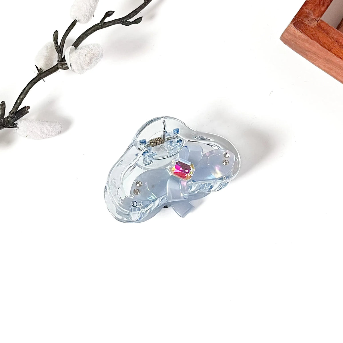 A light blue translucent hair claw clip with a matching ribbon bow and a pink iridescent rhinestone, displayed on a white background with decorative props.