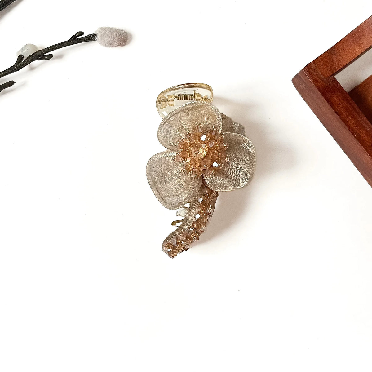 A gold-toned banana clip hair accessory decorated with a sheer champagne beige fabric flower and a textured metallic gold center, shown on a white background with decorative props.