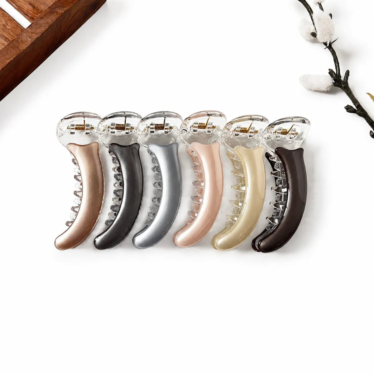 Six claw banana clips in assorted metallic and neutral colors, arranged in a row on a clean white background with subtle decorative props in the corners.